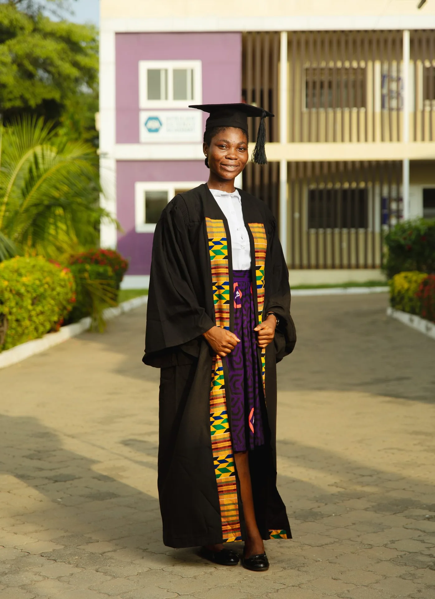 An ASA graduate in cap, gown and kente sash on campus